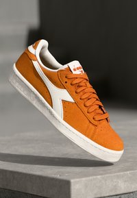 Orange suede sneaker with white accents, textured sole, and perforated detailing. Features flat orange laces and a branded tongue.