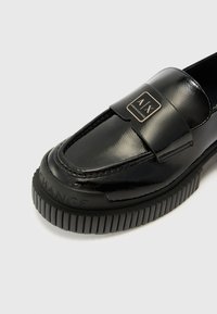 Juodos patentinės odos loafer'iai su plokščia, grioveliuota gumine padu. Yra ryškus logotipo ženkliukas ant dirželio ir dekoratyvinė siuvinėta detalė.