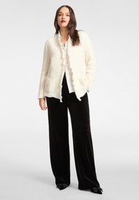 Open-front crèmekleurig jack met rafelige randen, textuurstof; gecombineerd met een lichte blouse en zwarte broek met wijde pijpen.