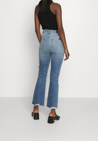 Jeans acampanados de cintura alta en denim azul medio, con dobladillo deshilachado, dos bolsillos traseros y una etiqueta en la cinturilla. Combinados con tacones negros.