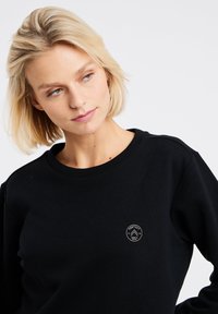Zwarte sweatshirt met ribbelboord bij de hals, voorzien van een klein cirkelvormig logo op de borst. De stof lijkt glad en duurzaam.