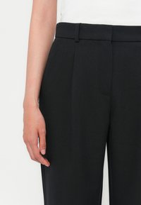 Sorte skreddersydde bukser med en jevn tekstur, flat front og beltehemper. Viser en finger som hviler på stoffet, som viser nærbilder av designdetaljer.