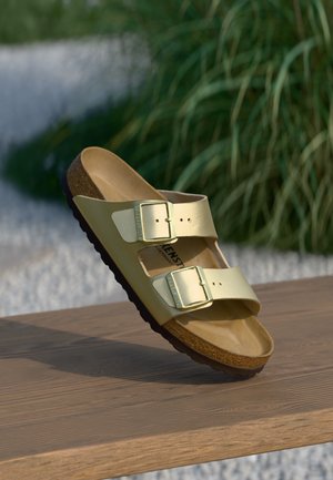 Metaliczne złote sandały Birkenstock z dwoma regulowanymi klamrami, spoczywające na drewnianej powierzchni, w tle rozmyte zielone rośliny.