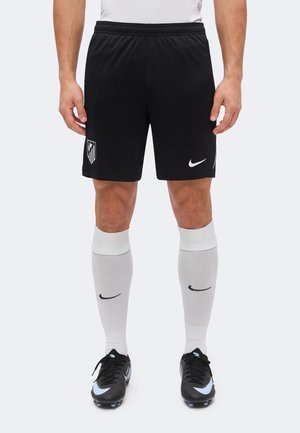 Muž nosiace čierne športové šortky s logom športového tímu a emblémom Nike, biele podkolienky a čierne Nike futbalové kopačky.