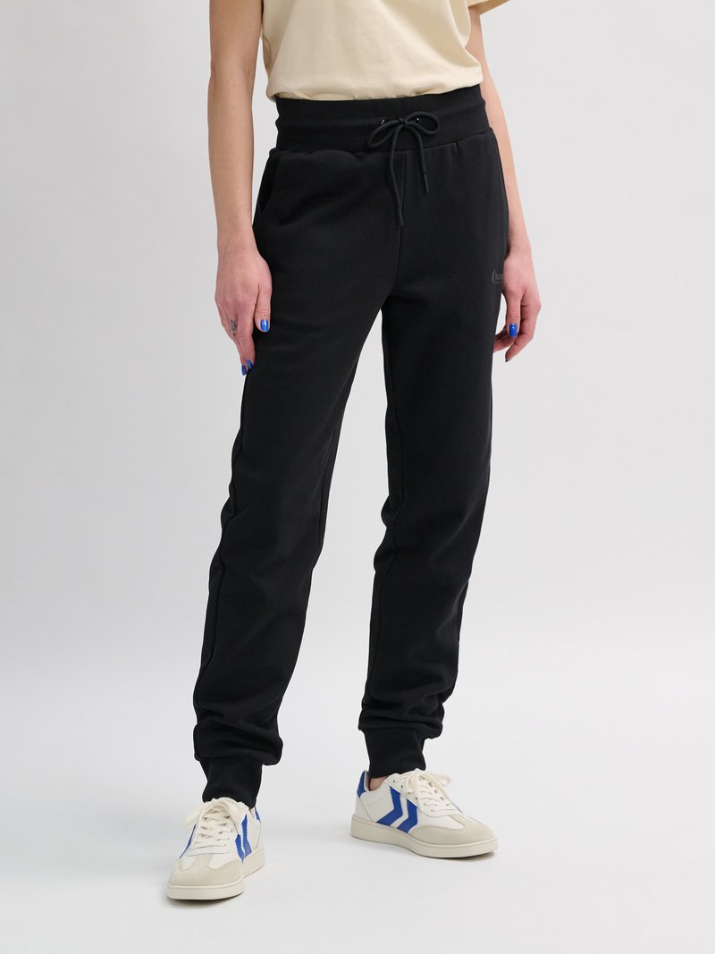 Person, die schwarze Sweatpants mit Kordelzug, beige-blaue Sneakers und ein beiges kurzärmeliges Oberteil trägt, steht vor einem einfarbigen Hintergrund.