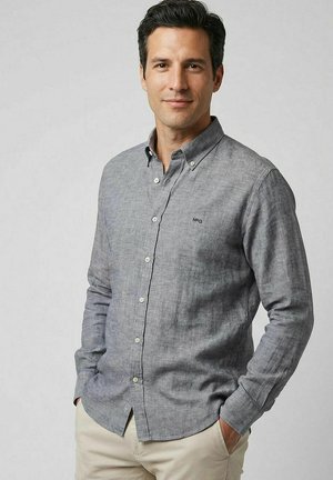 Man met donker haar, die een lichtgrijs overhemd met knopen en beige broek draagt, staand met handen in de zakken tegen een effen achtergrond.