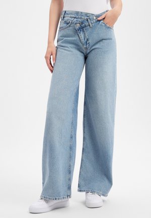 Hellblaue Jeans mit weitem Bein und asymmetrischem Knopfverschluss, getragen mit weißen Sneakers und weißem Oberteil, Hand ruht in der Tasche.