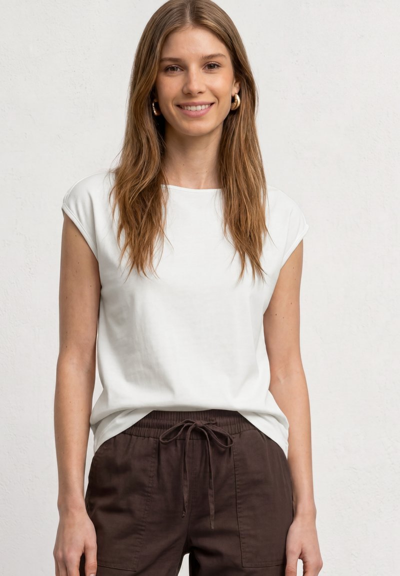 Jeune femme aux longs cheveux bruns souriante, portant un haut blanc sans manches et un pantalon marron foncé à cordon sur un fond clair uni.