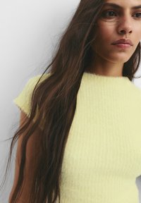 Fittelt sárga kötött felső rövid ujjakkal, textúrált anyagból, kerek nyakkivágással, hosszú sötét hajjal kiemelve.
