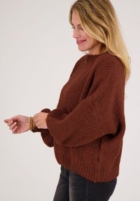 Vrouw die gouden armbanden rechtzet, draagt een losse, dikke bruine trui en donkere spijkerbroek tegen een effen witte achtergrond.