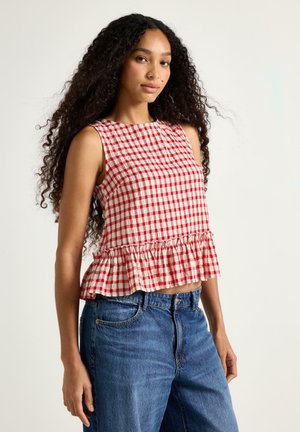 Kvinde med langt krøllet hår iført ærmeløs rød og hvid ternet top med flæsekant og blå jeans, stående mod en ensfarvet baggrund.