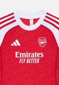 Maillot de football rouge avec un motif texturé, des accents blancs et trois bandes sur les épaules. Affiche le logo d'Arsenal et le texte du sponsor.