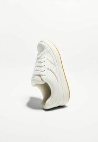 Fehér sneaker sima bőr felsőrésszel, kerek orral, lapos gumitalppal és textúrált nyelvvel, ami fűzős kialakítást és diszkrét oldalsó díszítéseket tartalmaz.