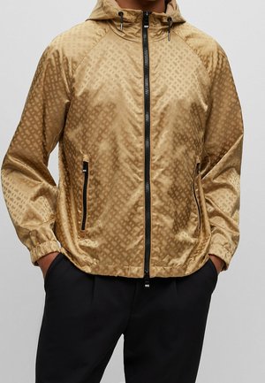 Man draagt een lichtbruine, met patroon bedrukte capuchonjas met zwarte rits en ritszakken, handen in de zakken van een zwarte broek.