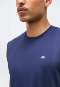 Κοντινό πλάνο ενός άνδρα που φοράει μπλε ναυτικό αμάνικο αθλητικό μπλουζάκι με μικρό λευκό λογότυπο στο στήθος μπροστά σε απλό φόντο.