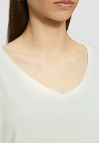 Biała koszulka polo z dekoltem w serek, o miękkiej, gładkiej fakturze, prezentująca luźny krój. Dekolt delikatnie zakrzywia się z czystym, stonowanym przeszyciem.