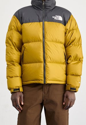 Mann trägt eine gepolsterte The North Face Jacke in Gelb und Schwarz mit Reißverschluss und braune Hose, steht vor einem schlichten Hintergrund.
