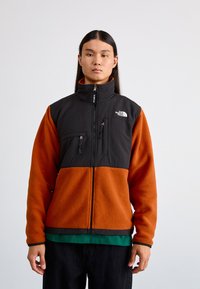 Giacca in fleece con una sezione superiore nera e una sezione inferiore arancione. Presenta un colletto alto, tasche frontali e chiusura con zip.