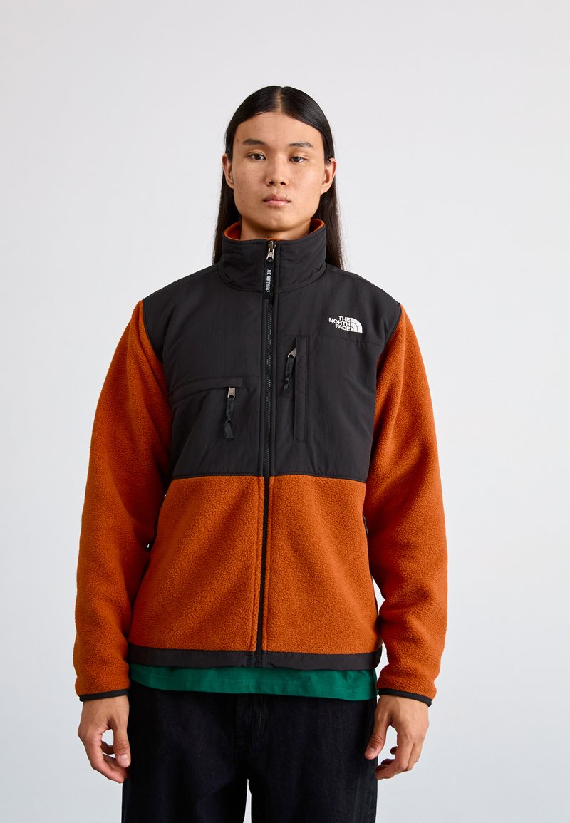 Giacca in fleece con una sezione superiore nera e una sezione inferiore arancione. Presenta un colletto alto, tasche frontali e chiusura con zip.