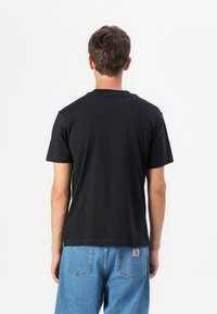 T-shirt en coton noir avec un col rond, des manches courtes et une coupe droite. Coutures visibles à l'ourlet et aux épaules, porté avec un jean bleu.