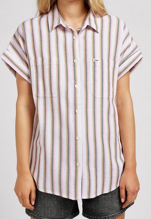 Femme portant une chemise à manches courtes boutonnée avec des rayures verticales roses, blanches, grises et jaunes et des poches poitrine, associée à un short foncé.