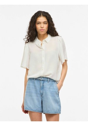 Femme aux longs cheveux bouclés portant une chemise blanche transparente à manches courtes et des shorts en denim bleu clair taille haute.