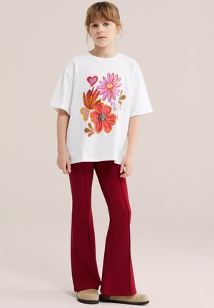 Otrok, ki nosi belo majico s pisanim potiskom cvetov in src, skupaj z rdečimi zvončastimi hlačami in bež čevlji, stoji na nevtralnem ozadju.