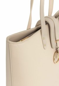 Beżowa skórzana torba na ramię z podwójnymi uchwytami, gładką teksturą i złotym metalowym akcentem w formie pierścienia. Otwarte wzornictwo, przestronne wnętrze widoczne.