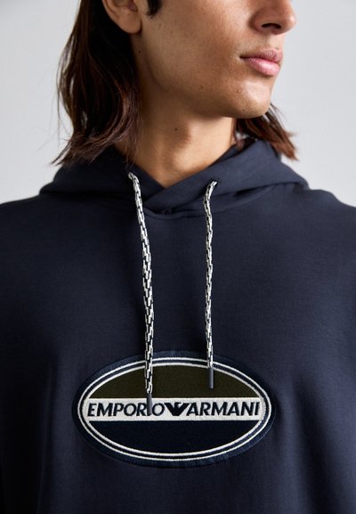 Tengerészeti kapucnis pulóver nagy, kör alakú Emporio Armani logóval. Kontrasztos fehér és fekete zsinórokkal rendelkezik, texturált felülettel.