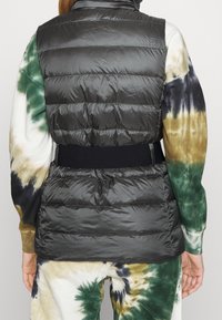 Fodrad väst i mörkgrå nylon med belagd midja, med horisontella quiltade sektioner. Bärs över en tie-dye skjorta med gröna och bruna nyanser.