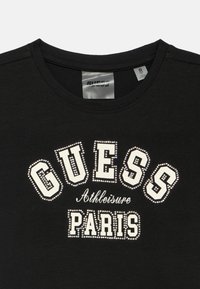 Μαύρο μπλουζάκι με στρογγυλή λαιμόκοψη, με λευκό κείμενο διακοσμημένο με στρας που γράφει "GUESS Athleisure PARIS" σε έντονη γραμματοσειρά. Μαλακό βαμβακερό υλικό.