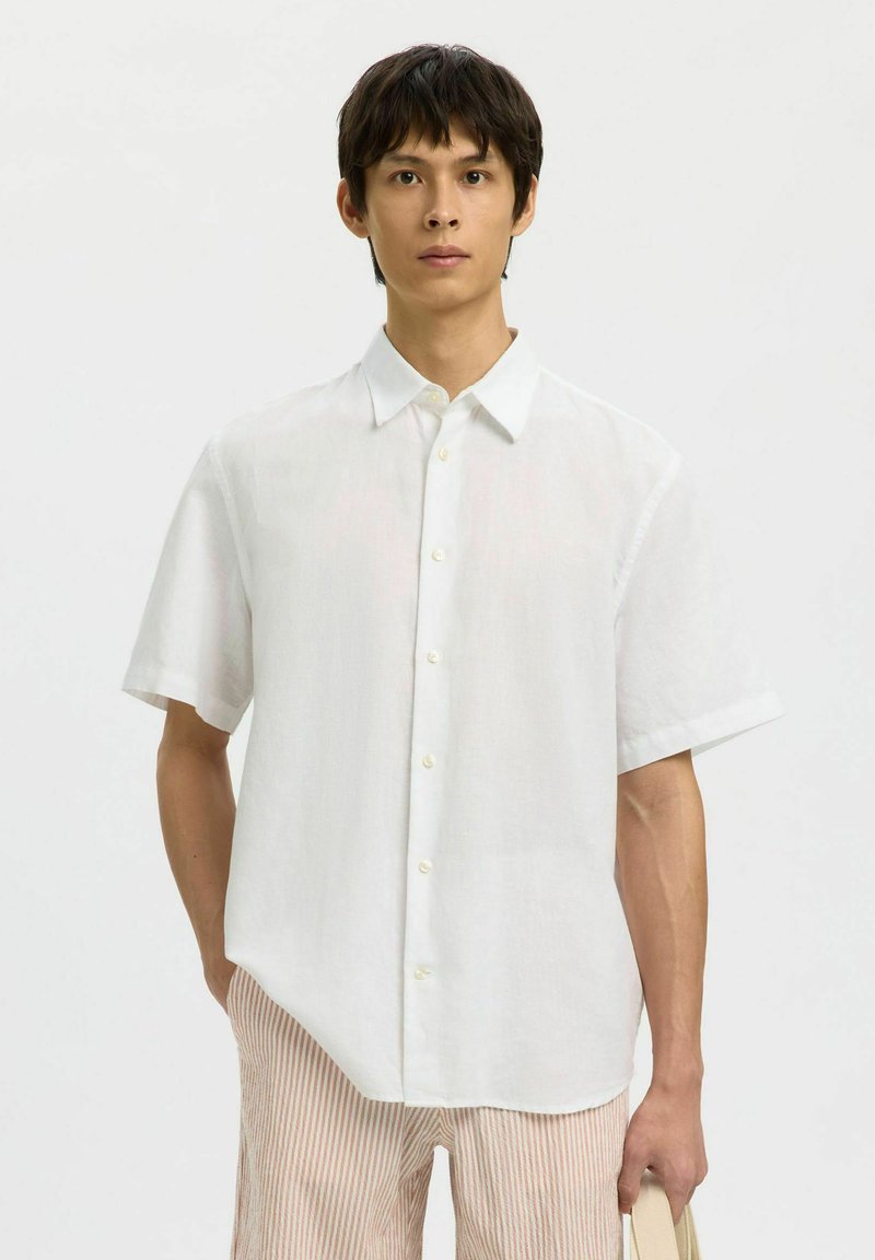 Homme portant une chemise blanche à manches courtes boutonnée et un pantalon rayé de couleur claire, debout devant un fond uni clair.