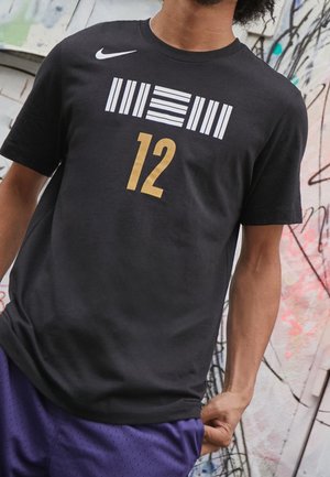 Persona con camiseta negra de Nike con barras geométricas blancas y número 12 en dorado, de pie junto a una pared con grafiti, sosteniendo unos pantalones cortos morados.