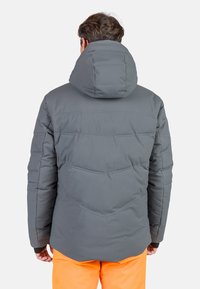 Graue Puffjacke mit Kapuze, die über gesteppten Details und einer leicht glänzenden Außenseite verfügt, getragen über leuchtend orangefarbenen Hosen.