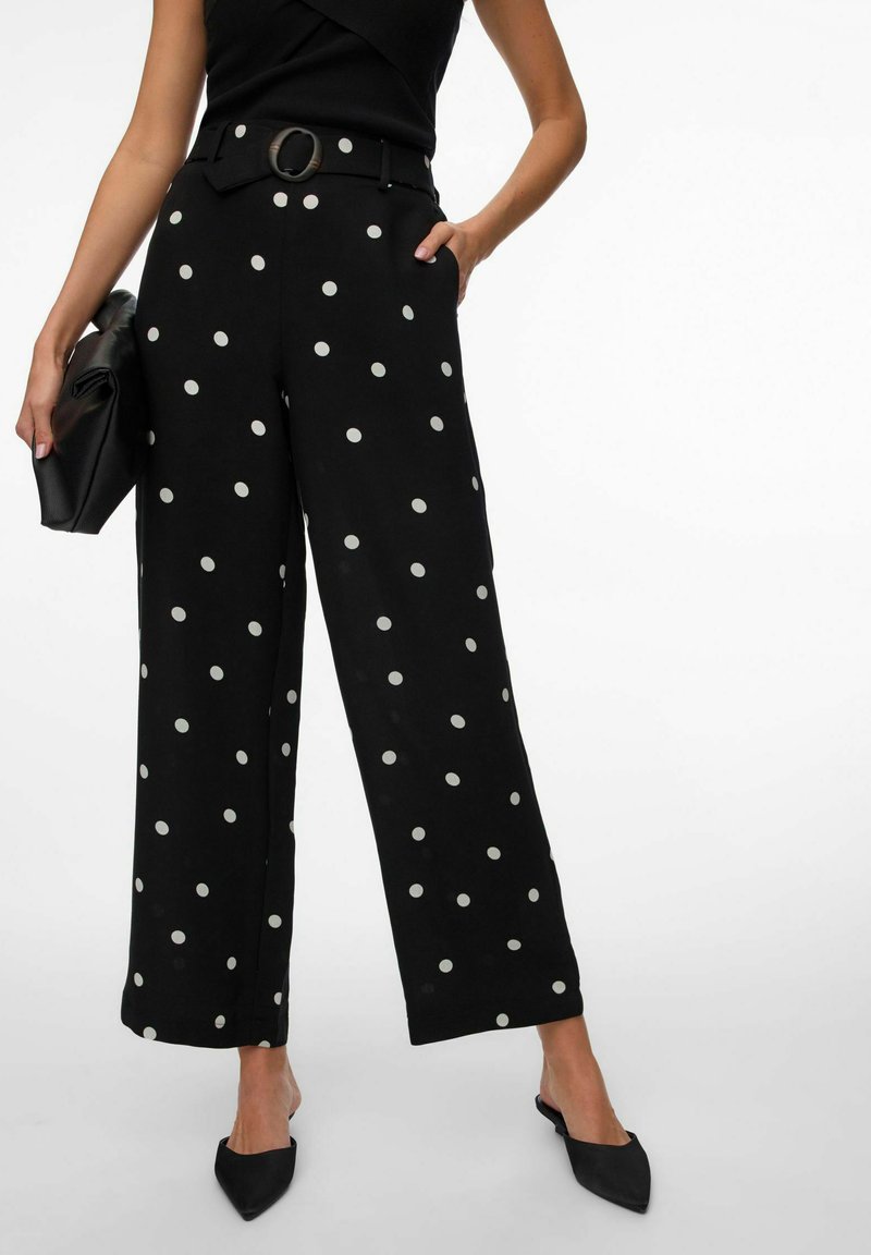 Femme portant un pantalon large noir à pois blancs, ceinture à la taille, chaussures plates noires pointues, et tenant une pochette noire.