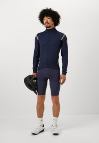 Giacca da ciclismo blu con accenti grigi, abbinata a pantaloni corti scuri con un motivo testurizzato. La persona tiene un casco nero e indossa sneakers bianche.