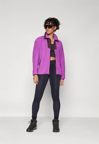 Osoba ubrana w za dużą fioletową kurtkę, czarny stanik sportowy, czarne legginsy, buty trekkingowe i duże czarne okulary przeciwsłoneczne, stojąca na tle bez wzorów.