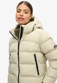 Giacca a vento beige con cappuccio, dotata di zip nere e colletto verde oliva texture. Include una patch con logo sulla spalla sinistra.