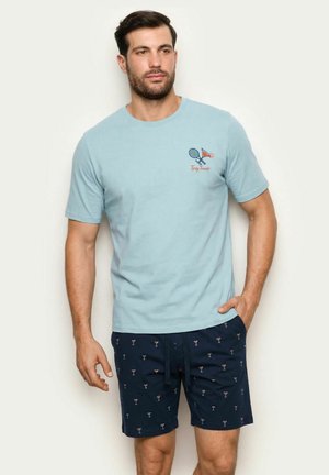 Hombre con camiseta azul claro con raquetas de tenis y texto "Tipsy Tennis" y pantalones cortos azul marino con patrón de pequeños vasos de cóctel.