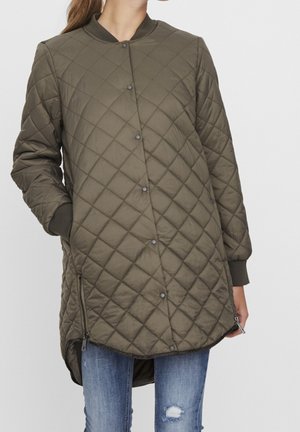 Žena má na sobě dlouhý olivově zelený prošívaný kabát se zapínáním na patentky, zipy na bocích a žebrované manžety, v kombinaci s potrhanými modrými džínami.