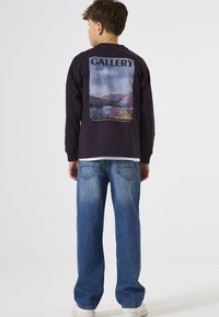 Donker shirt met lange mouwen en een grafische afdruk op de achterkant met bergen en water, gelabeld "GALLERY." Gepaard met lichtblauwe recht-toe-recht-aan spijkerbroek en witte sneakers.