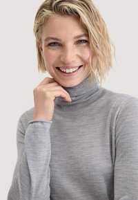 Lächelnde Frau mit kurzem, welligem, blonden Haar, die einen hellgrauen Rollkragenpullover trägt und ihre Hand entspannt nahe ihrem Kinn gegen einen einfarbigen Hintergrund stützt.