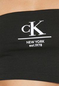 Černý bandeau top vyrobený z elastické látky, s bílým logem, které zní "cK NEW YORK est. 1978" umístěným na přední straně. Hladký povrch.