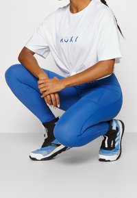 Leggings atléticos azules con líneas de costura, acompañados de una camiseta blanca holgada con la marca "ROXY" y calzado negro con detalles azules.
