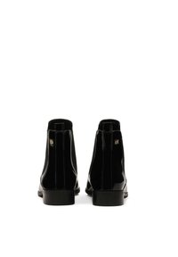 Botines de charol negro con una textura suave, puntera redondeada y tacón plano, que presentan un sutil acento de logo cerca de la cremallera.