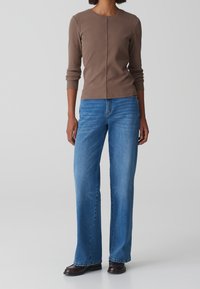 Kvinna iförd en långärmad brun topp, blå vida jeans och mörkbruna skor som står mot en enkel ljus bakgrund.