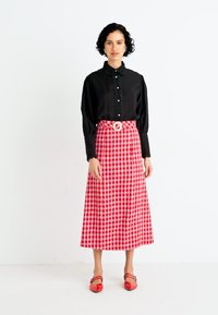 Camisa negra de manga larga con volantes texturizados, combinada con una falda maxi a cuadros rojos y blancos y unos zapatos planos rojos con un detalle de hebilla.