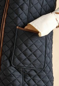 Kvistīts tumši zils veste ar brūnu apmali un priekšējo kabatu. Audums ir gluds, ar dimanta formas šuvēm.