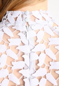 Valkoinen pitsitoppi, jossa on kukkakuosi, läpinäkyvä verkkoalusta, takana vetoketju ja kaksi kankaalla päällystettyä nappia kauluksessa.