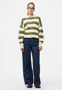 Črtast pleteni kardigan v olivni in krem barvi, kratkega kroja, z gumbi. Kombiniran z temnimi kavbojkami z visokim pasom in širokimi nogavicami. Črni čevlji.